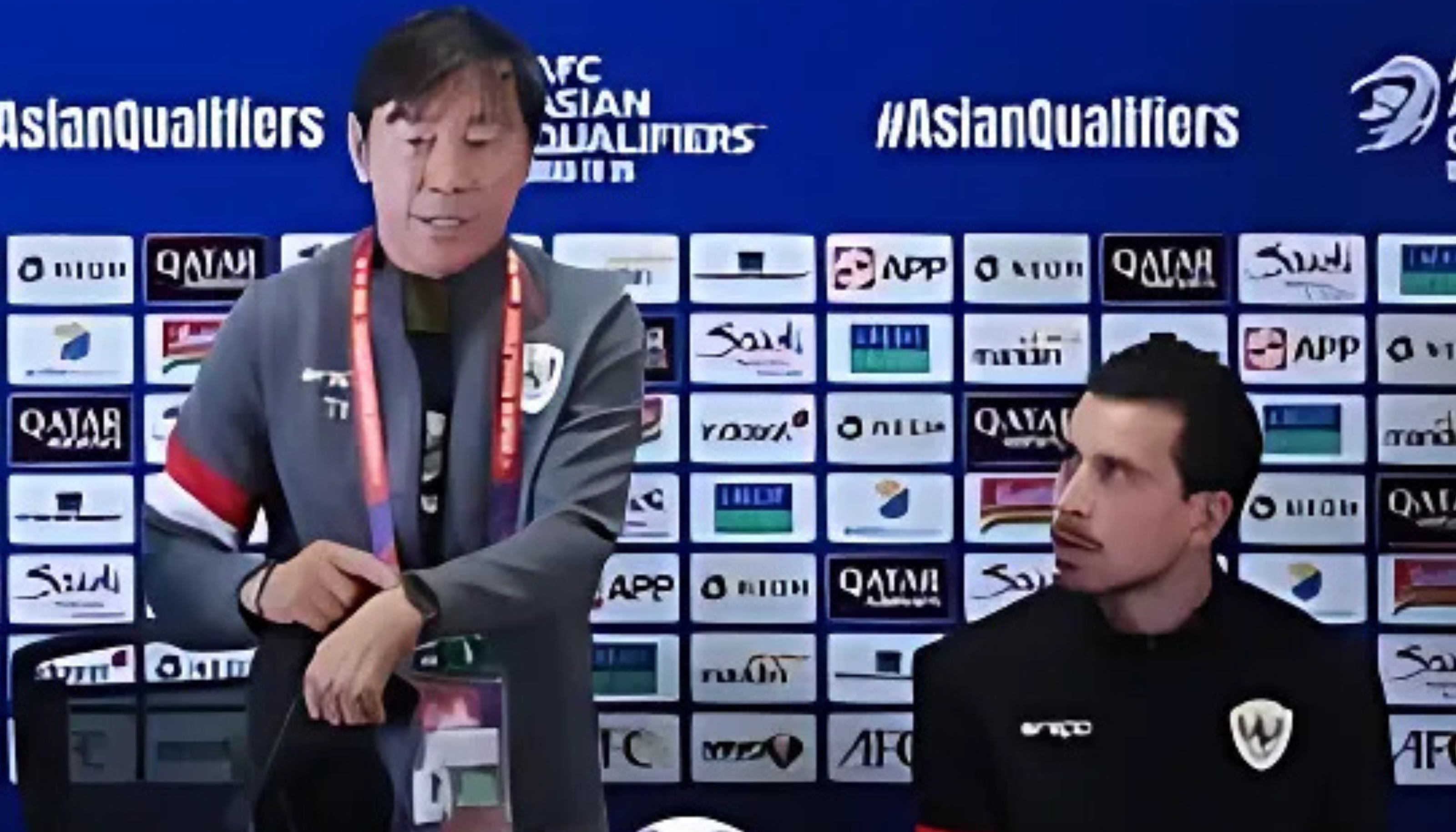Shin Tae-yong sedang berbicara dalam konferensi pers bersama seorang pemain, dengan latar belakang logo sponsor