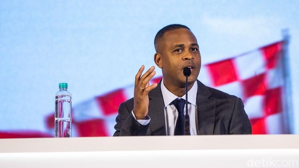 Patrick Kluivert berdiri di depan logo klub sepak bola dengan mengenakan setelan jas biru sambil tersenyum