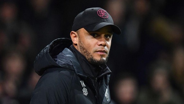Vincent Kompany sedang berdiri di pinggir lapangan dengan ekspresi serius, mengenakan jaket hitam dan topi.