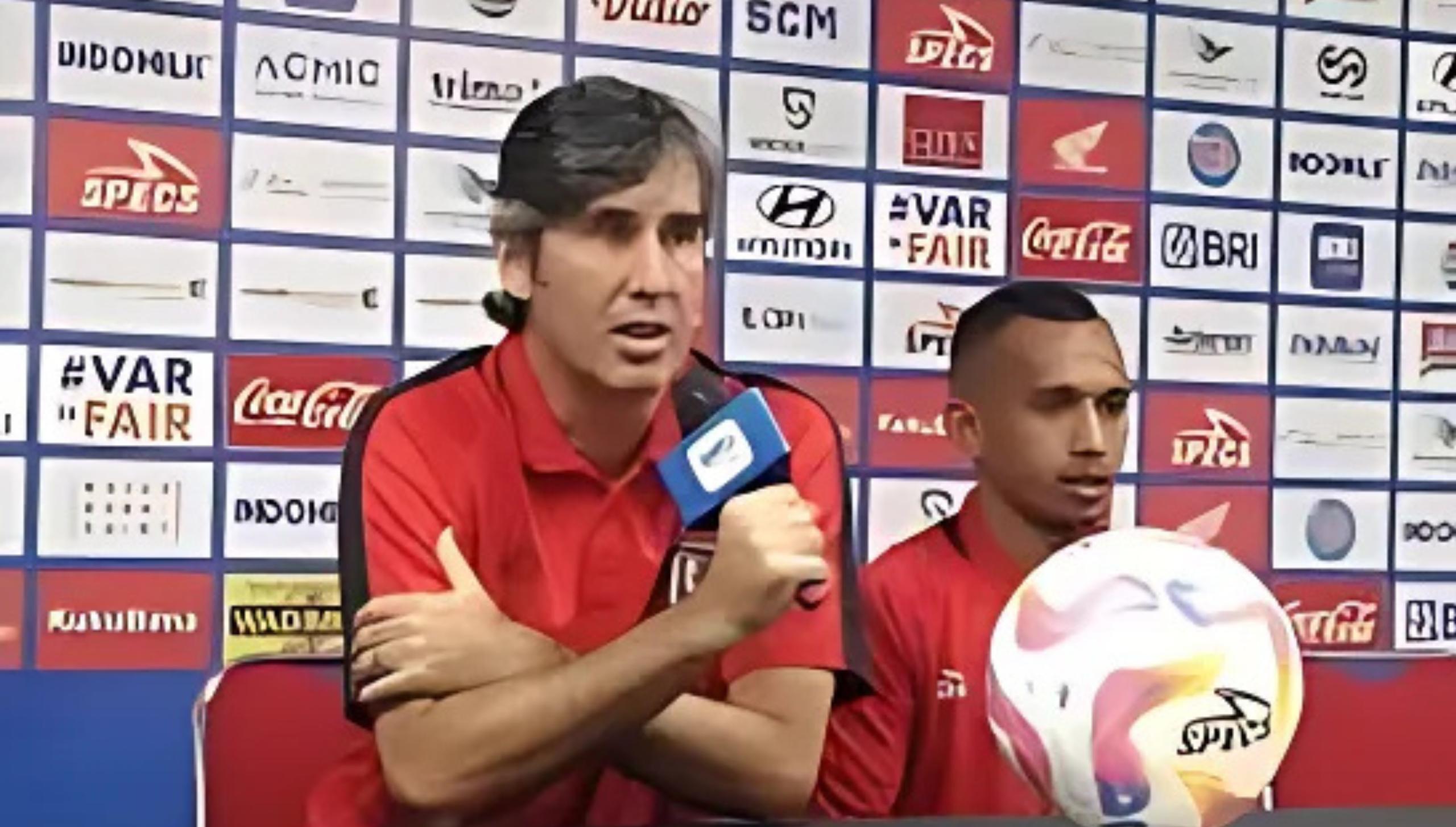 Pelatih Bali United berbicara dalam konferensi pers didampingi seorang pemain dengan latar belakang papan sponsor
