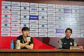 Empat pemain lini tengah Timnas Indonesia dalam aksi, dengan siluet Shin Tae-yong di atasnya dan logo Piala Dunia 2026 di sudut kanan. Latar belakang gelap dengan sorotan cahaya emas.